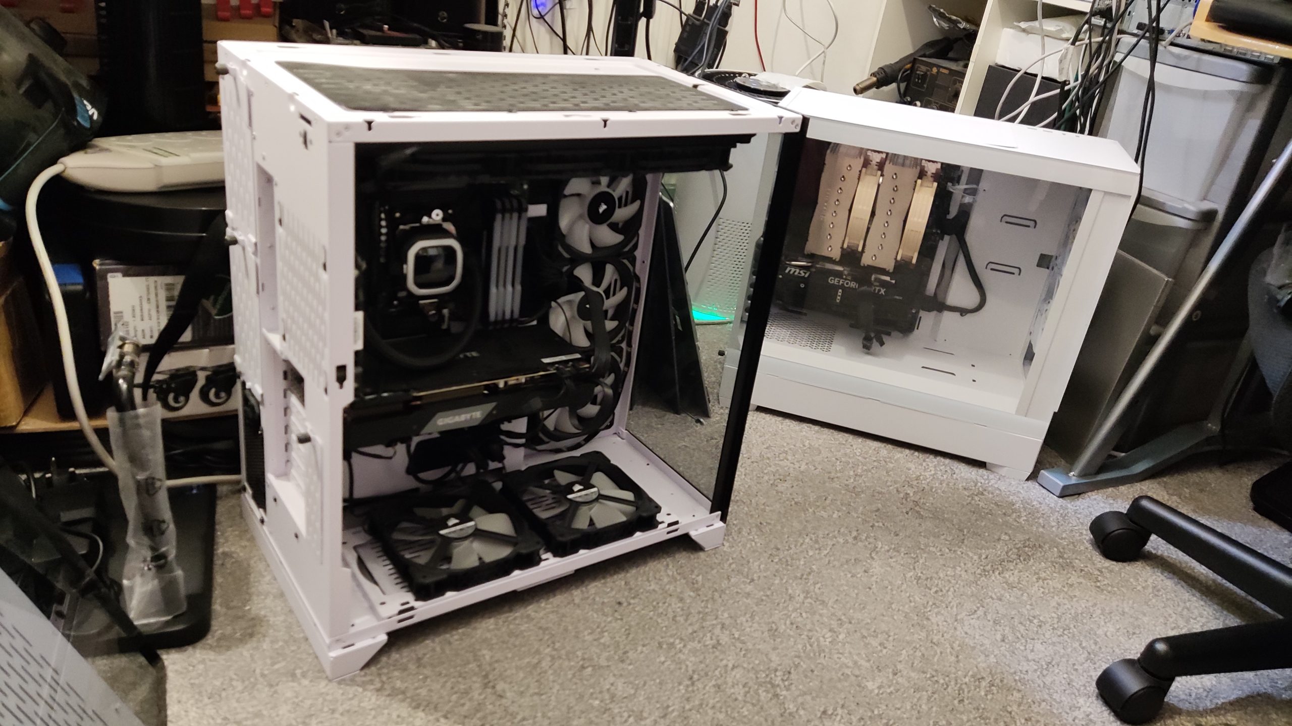 Computer repair workstation showing a white desktop computer case with both side panels removed, positioned on a grey carpeted floor. The open case reveals internal components including black cooling fans, a graphics card with "GEFORCE" branding visible, motherboard, and various connecting cables in black and coloured wires. The computer case appears to be a mid-tower design with a mesh front panel and is currently being serviced or assembled. In the background, electronic equipment and components are visible on white shelving units and work surfaces, including what appears to be additional computer hardware and repair tools. A black office chair on wheels is positioned in the lower right corner of the frame. The setting appears to be a professional computer repair workshop with adequate lighting, demonstrating active computer repair and diagnostic services in progress. The opened computer case shows the systematic disassembly typical of professional computer repair work, with components accessible for diagnosis, replacement, or upgrade procedures.