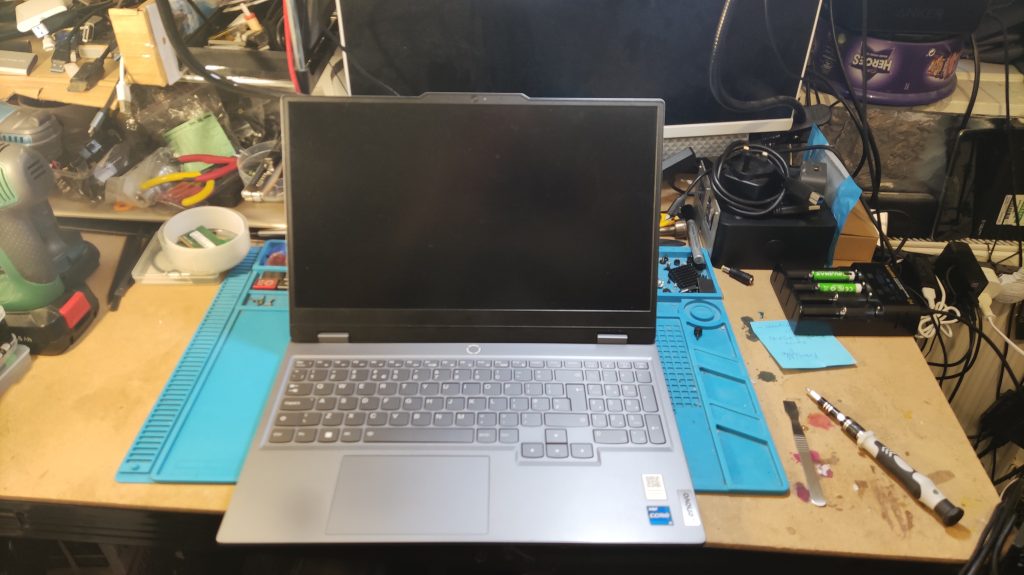 Silver laptop computer positioned on a blue anti-static repair mat within a professional computer repair workshop. The laptop appears to be a modern model with a full-size keyboard including numeric keypad and large trackpad. The screen is currently black or powered off. The device is placed centrally on the blue mat which features organized compartments and measurement rulers. Surrounding the workspace are various repair tools and equipment including white tape dispensers, coloured markers, precision screwdrivers, and electronic testing equipment visible in the background. Additional computer components and cables are organized on wooden shelving units behind the workstation. The setup demonstrates active laptop repair services with proper anti-static precautions and professional tool organization typical of expert computer repair facilities.
